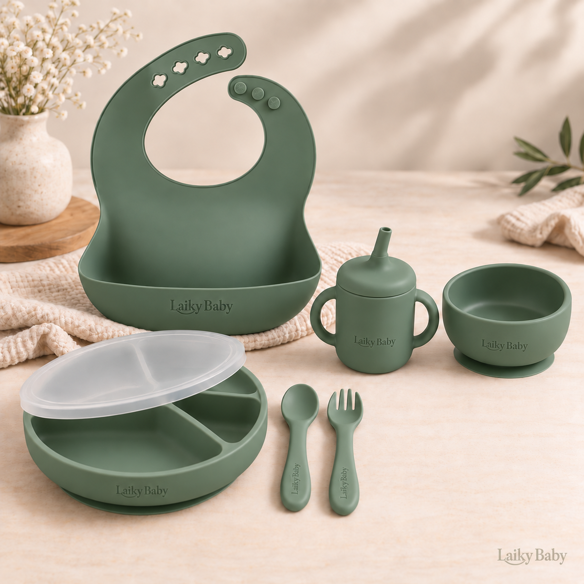 A coordinated silicone baby feeding set in muted sage green, including a large silicone bib, sippy cup, small bowl, plate with lid, spoon, and fork. Comes in Blush, Sage, Ether and Shifting Sand