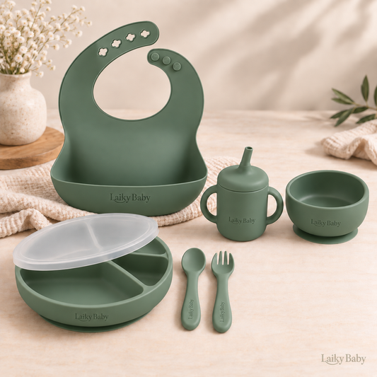 A coordinated silicone baby feeding set in muted sage green, including a large silicone bib, sippy cup, small bowl, plate with lid, spoon, and fork. Comes in Blush, Sage, Ether and Shifting Sand